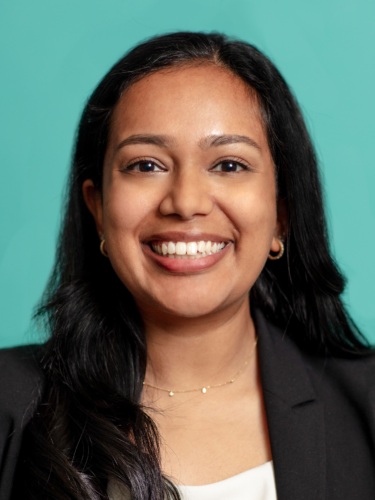 A person with long black hair smiles, dressed in a black blazer and white top, with a turquoise background.